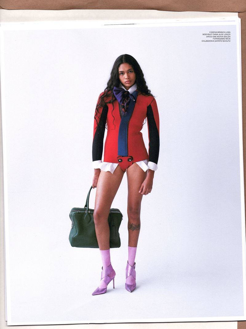 A young woman poses in a red jacket with black sleeves, a blue bow, white shirt cuffs, briefs, pink socks, and lilac heels, holding a green bag.