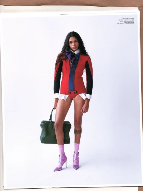 A young woman poses in a red jacket with black sleeves, a blue bow, white shirt cuffs, briefs, pink socks, and lilac heels, holding a green bag.