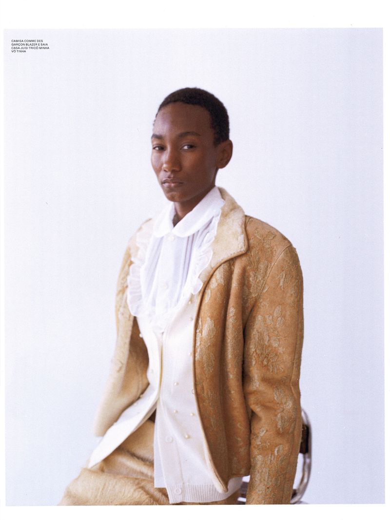 An adult female model with short dark hair poses in a golden-brown patterned suit with a white ruffled blouse against a white backdrop.