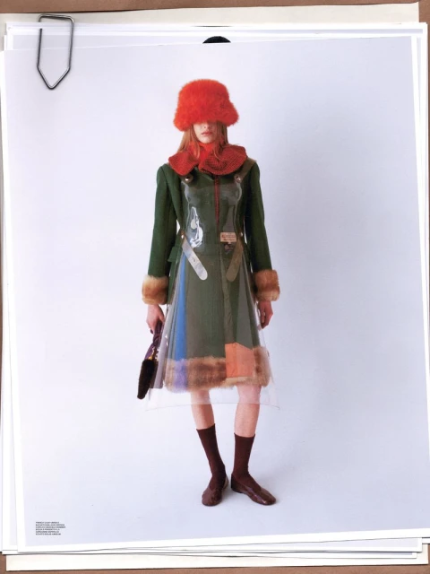 A fashion shot shows a model in a green jacket and clear dress, posing against a white backdrop with warm, natural lighting and a unique style.