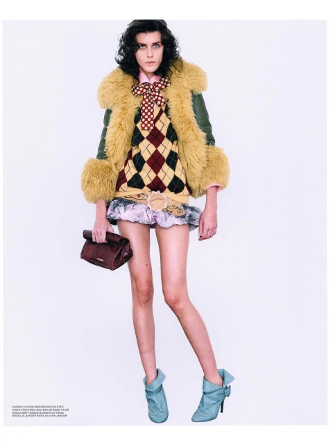 Full-length studio shot of a female model wearing a faux fur trimmed coat, argyle sweater, mini skirt, heeled boots, and carrying a handbag.