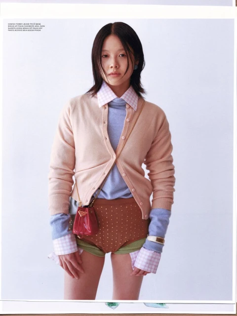 Asian woman in a layered fashion ensemble with a pink cardigan, brown shorts, and a red purse against a clean, white backdrop.