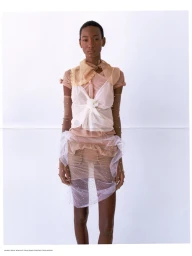 Model standing against a white backdrop wearing layered sheer organza, a knotted white top, and pearl-embellished sheer sleeves.