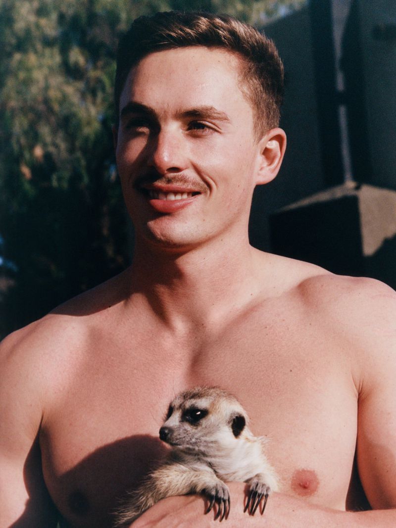 Bare-chested man holds a meerkat, facing the camera in an outdoor setting with dappled sunlight and a building in the background.