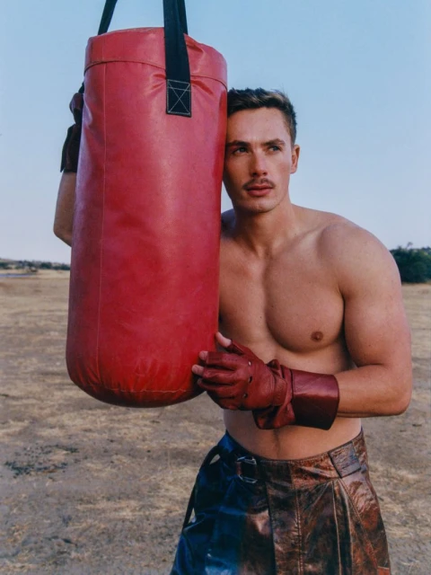 Shirtless male model poses with a red punching bag outdoors wearing brown leather skirt and boxing gloves under a clear sky.