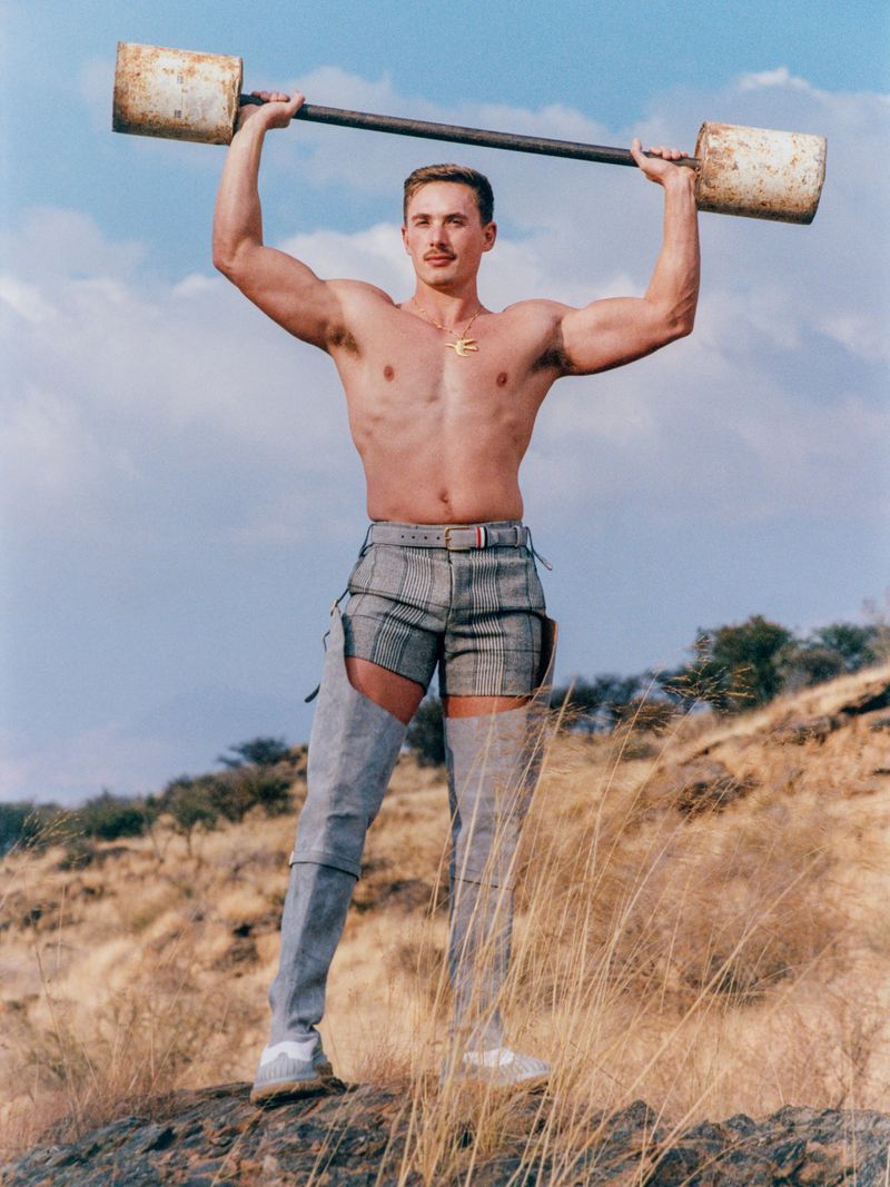 Muscular man outdoors lifts rusty barbell in avant-garde fashion, wearing shorts, chaps, and sneakers, set against a natural landscape.