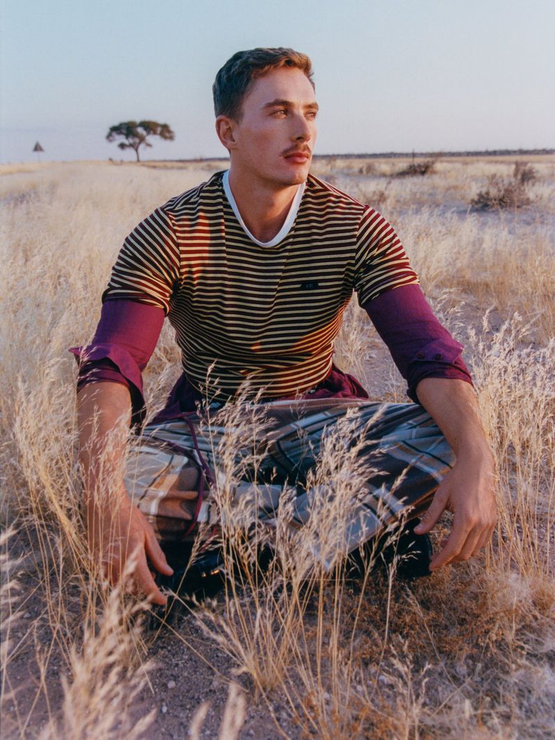 Man with mustache in striped shirt and plaid pants sitting in tall dry grass in a golden field with a distant tree in the background.