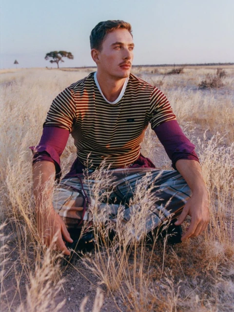Man with mustache in striped shirt and plaid pants sitting in tall dry grass in a golden field with a distant tree in the background.