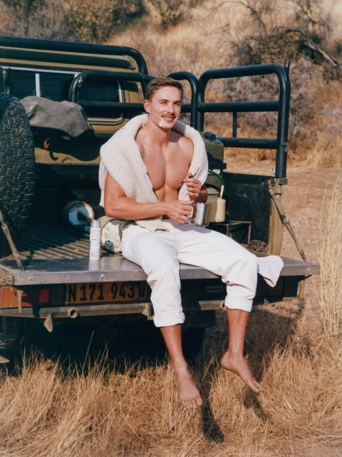 Shirtless man with shaving cream on face, sitting on a truck in a field. Tan vegetation and arid background. Beauty shot, outdoors.