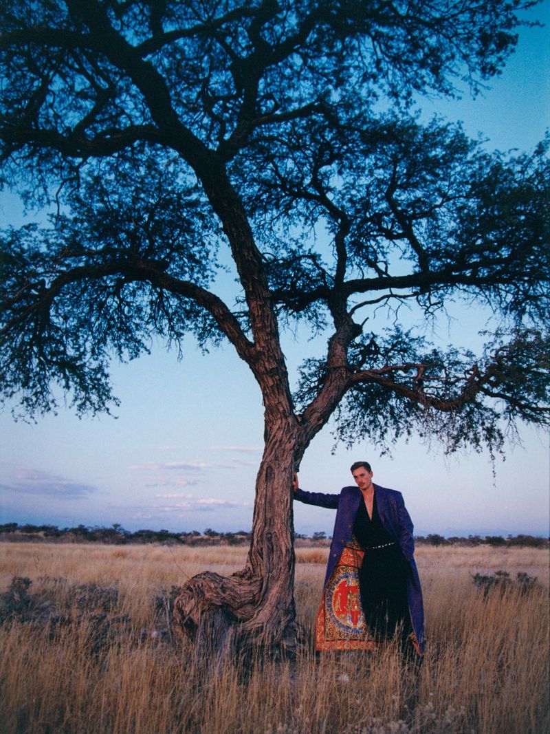 Man in long coat stands beside a tree in a field at dusk, featuring a twilight sky, dry grass, and sparse shrubs in the background.