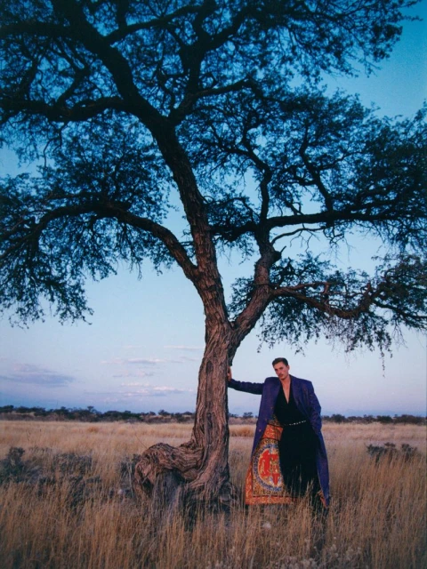 Man in long coat stands beside a tree in a field at dusk, featuring a twilight sky, dry grass, and sparse shrubs in the background.