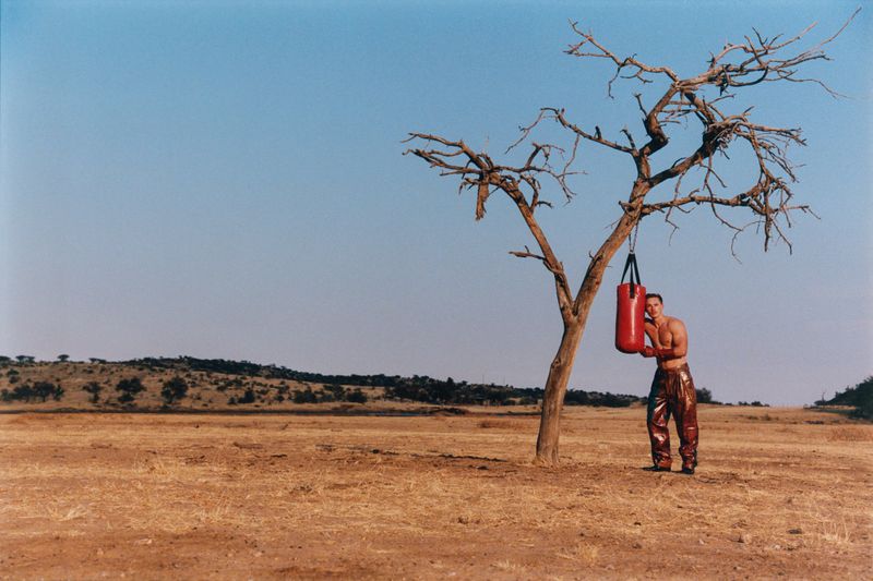 Adult man in athletic pants leaning against a red punching bag hanging from a bare tree in an arid, grassy landscape under a clear sky.