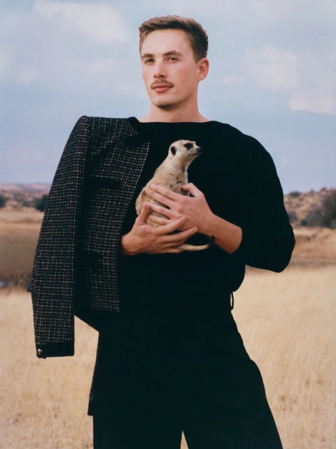 Adult male in a black top holds a meerkat in an outdoor setting, wearing a black and white blazer over his shoulder in natural light.