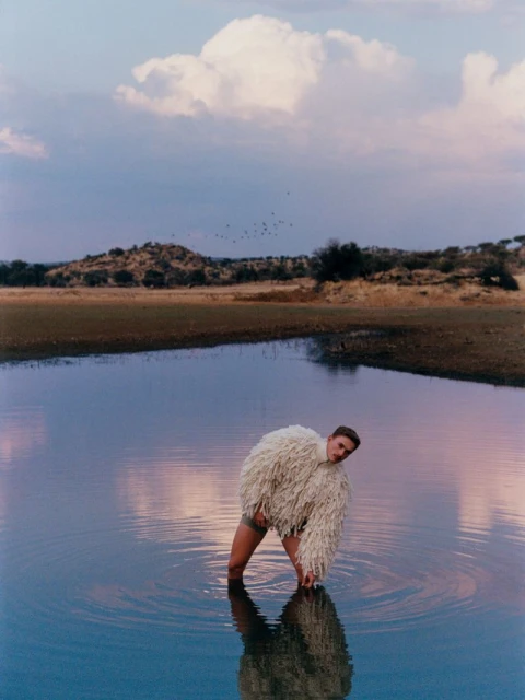 Adult male in a white, fringe jacket stands in a lake. Ripples surround him. Background landscape is comprised of cloudy sky, trees, & grass.