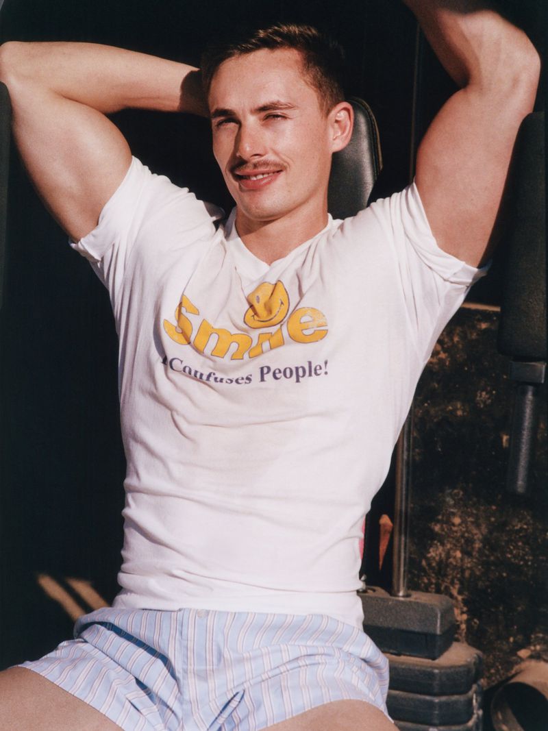 Man with muscular physique wearing a 'Smile Confuses People' t-shirt, seated in an indoor fitness setting, posing with his hands behind his head.