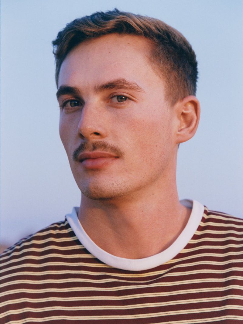 Portrait of a young adult male with short brown hair and a mustache, wearing a striped shirt against a soft blue background.