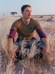 Man with mustache wearing a striped shirt and patterned trousers seated in a dry grassland landscape under an open sky.