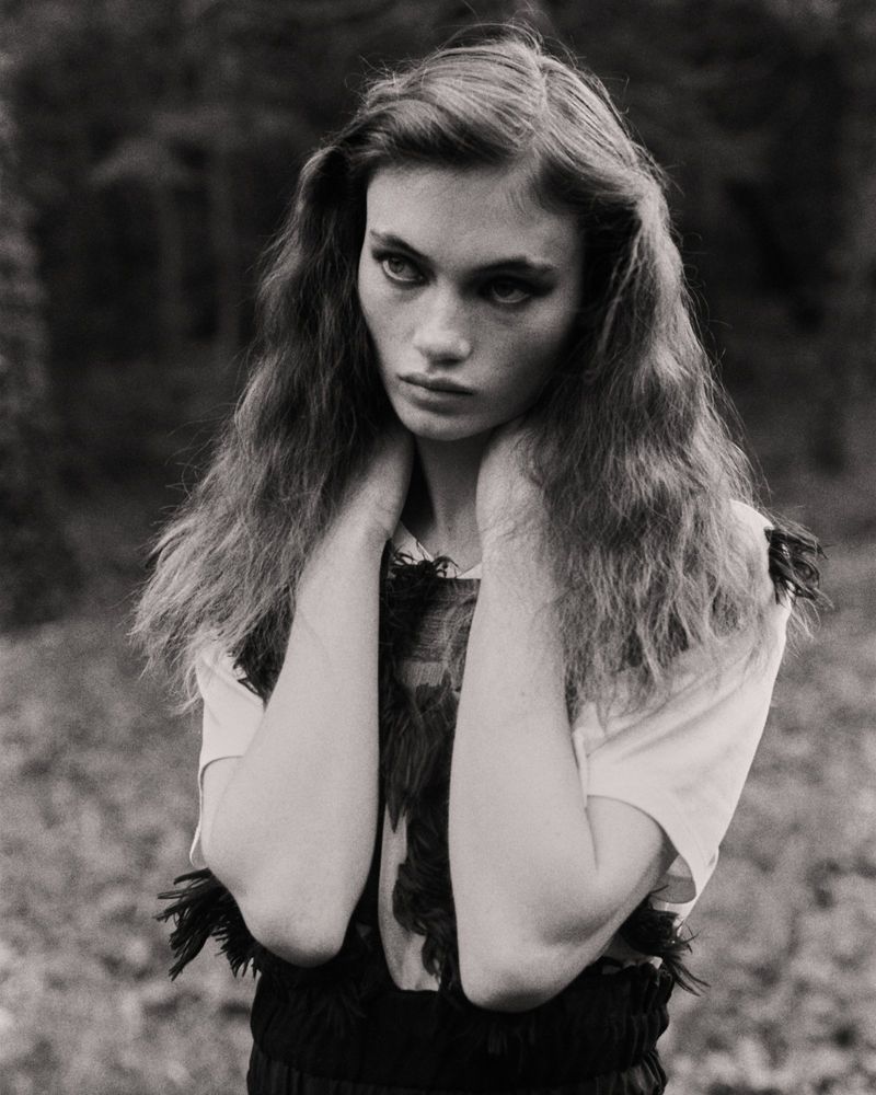 Monochrome portrait of a young woman with wavy brown hair and hands on her neck, looking off to the side in an outdoor setting.