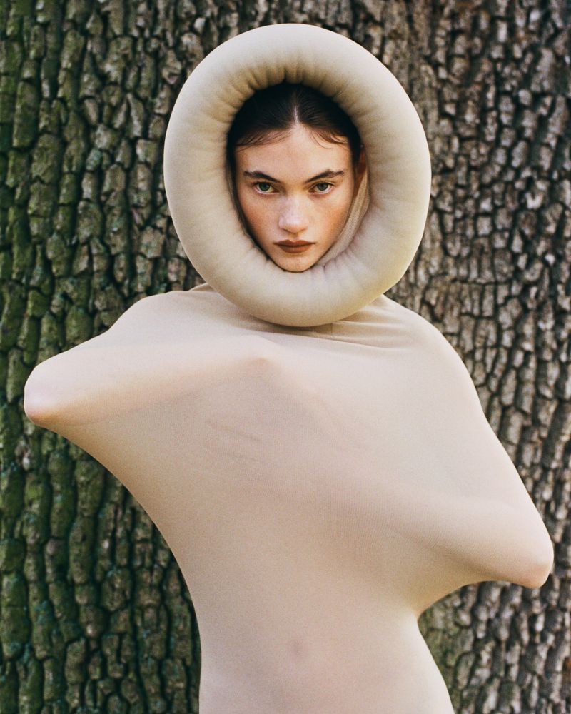 Woman in a beige dress and circular headpiece stands against a tree trunk background, featuring a combination of fashion and nature.