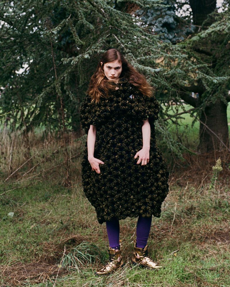 A young woman standing outdoors wearing a dark, textured dress with bright tights and gold boots, posing in a natural setting with trees.