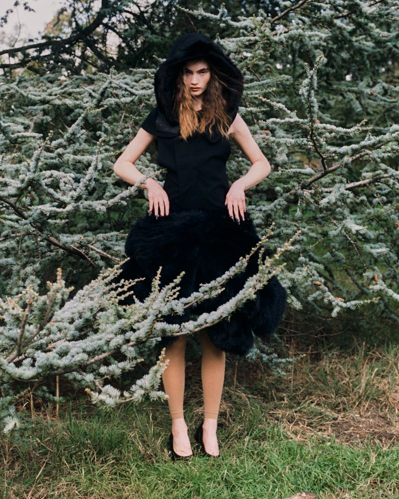 A fashion model poses in a dark dress with an oversized hood and textural lower skirt against a backdrop of green foliage.