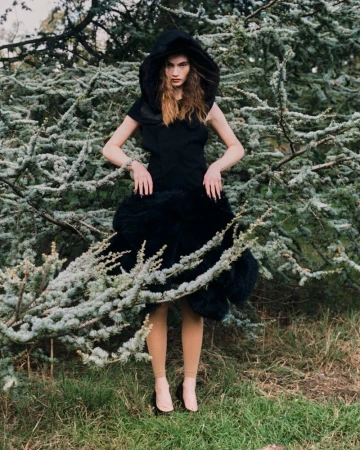 A fashion model poses in a dark dress with an oversized hood and textural lower skirt against a backdrop of green foliage.