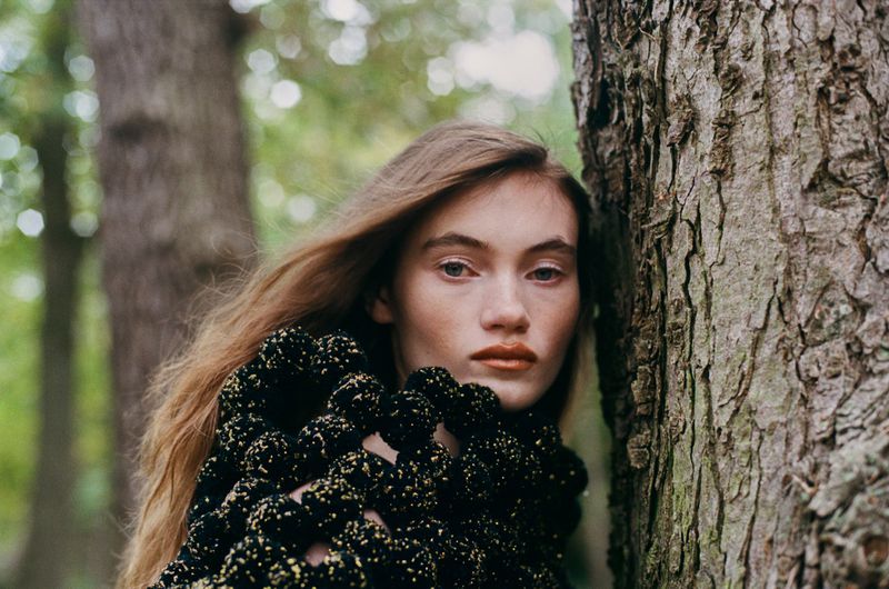 Portrait of a young woman with long brown hair in a patterned jacket standing next to a tree in an outdoor forest setting.
