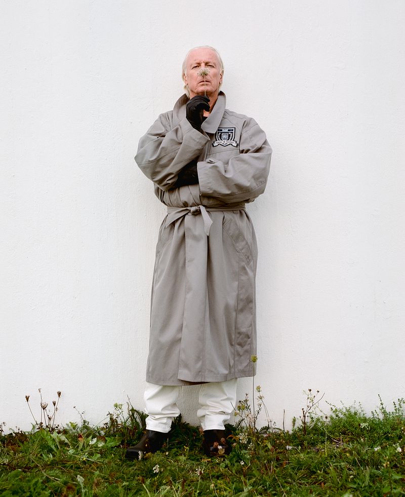 Full-length portrait of an older man with a mustache wearing a grey trench coat, white pants, and black gloves standing outdoors against a white wall.