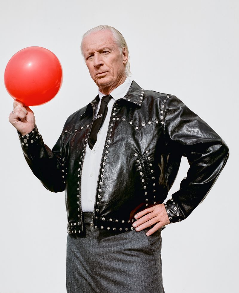 Older man in leather jacket poses with red balloon against a white backdrop, creating a striking contrast and playful yet serious mood.