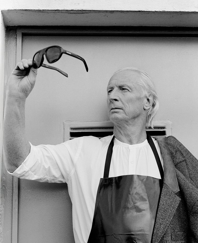 Elderly man in black-and-white portrait holding sunglasses, wearing an apron and jacket against a white wall with vent.