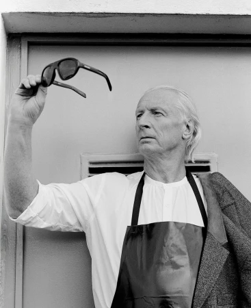Elderly man in black-and-white portrait holding sunglasses, wearing an apron and jacket against a white wall with vent.