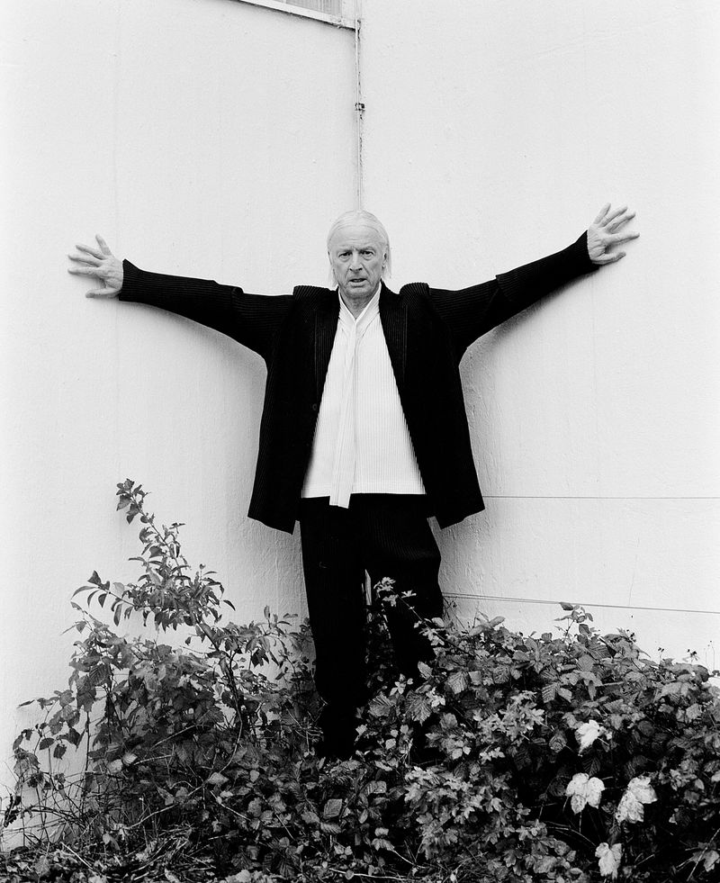 A man in a black suit coat poses with arms outstretched against a white wall, framed by greenery in a black and white photograph.