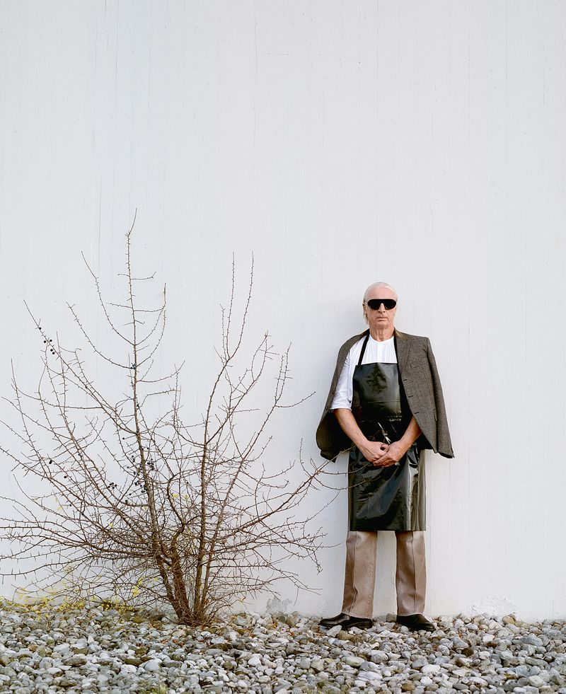 Full shot of an adult with sunglasses and apron, standing next to a bare bush against a white wall, with gravel on the ground.