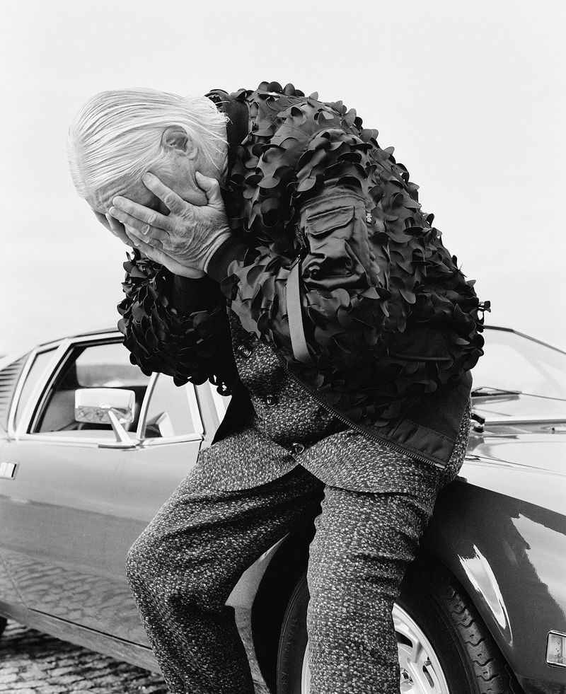 Elderly man in patterned suit and decorative bomber jacket leans against a car, face in hands, in a black-and-white studio shot.