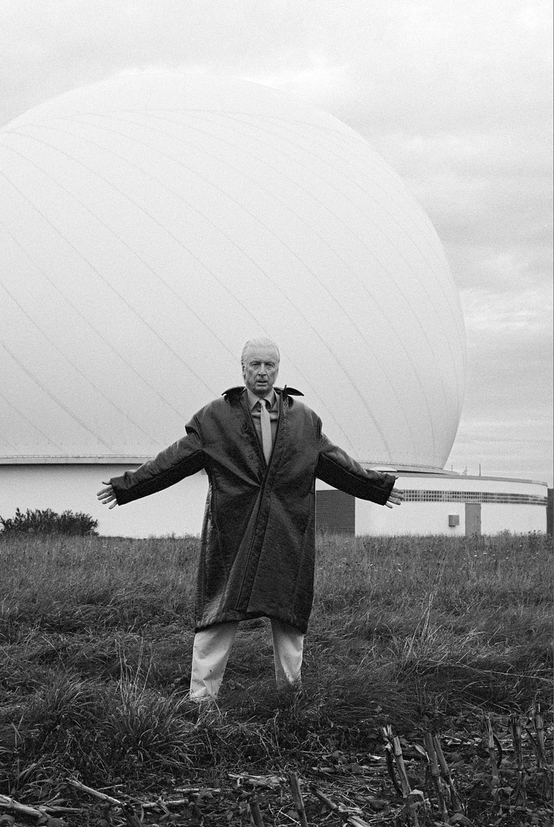 Man in oversized coat stands in grassy field before large geodesic dome building on cloudy day in black and white, arms outstretched.