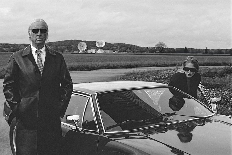 Older man in coat and sunglasses stands next to a car with woman in a black and white outdoor fashion shot near satellite dishes.