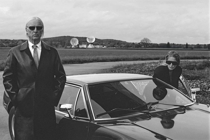 Older man in coat and sunglasses stands next to a car with woman in a black and white outdoor fashion shot near satellite dishes.