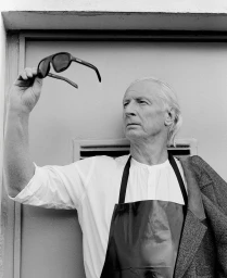 Black and white portrait of an older man holding up dark sunglasses while wearing a white shirt and black apron.