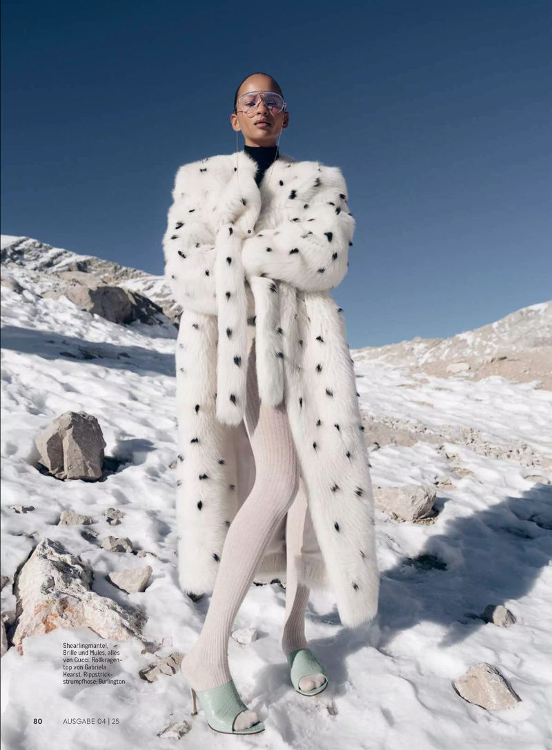 Woman in fashionable white faux fur coat poses on snowy, mountainous terrain under bright blue sky, wearing glasses and mint green mules.
