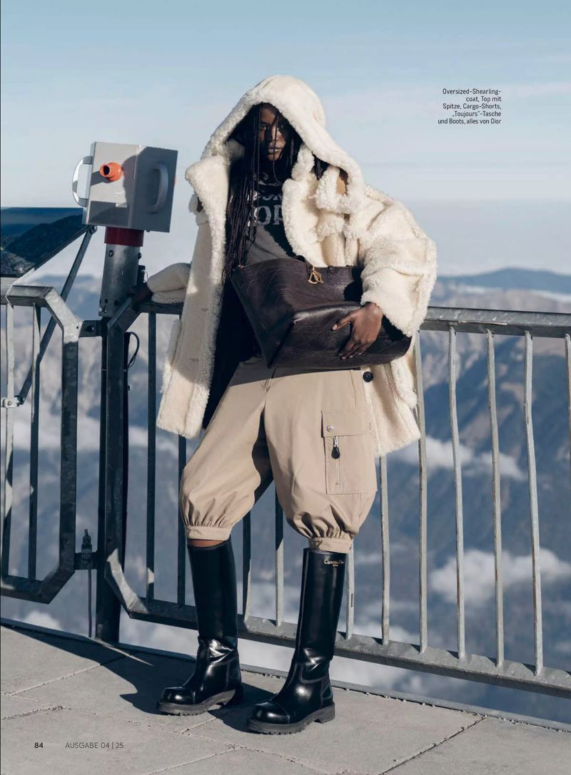 Fashion editorial shot of a Black female model wearing an oversized shearling jacket, cargo shorts, and black boots on a mountain top.