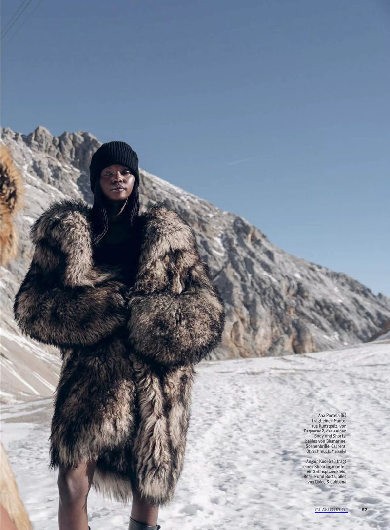 A woman in a furry coat and black knit cap stands in a snowy mountain setting on a bright, sunny day in a full outdoor shot.