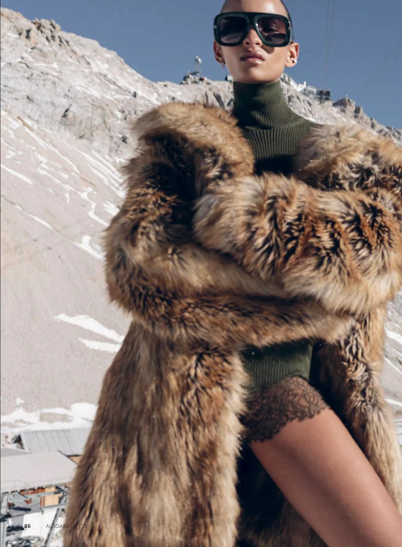 Adult female model wearing fur coat and sunglasses poses in a snowy alpine setting with mountains in the background on a sunny day.