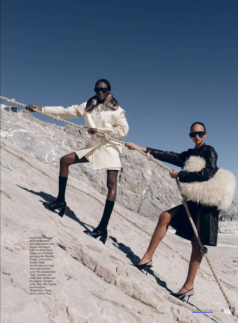 Two Black women posing on rocky terrain, wearing designer coats and sunglasses, holding a rope, against a clear blue sky. Fashion editorial.
