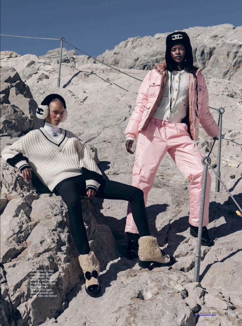 Two women pose in Chanel skiwear on a rocky mountain with blue sky. One sits while the other stands near a metal safety railing.
