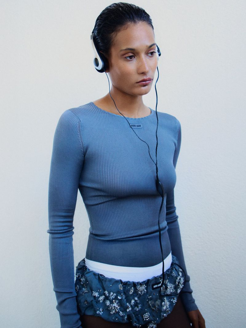 Woman wearing headphones and a blue ribbed shirt poses in front of a white wall in a studio environment with overhead lighting.