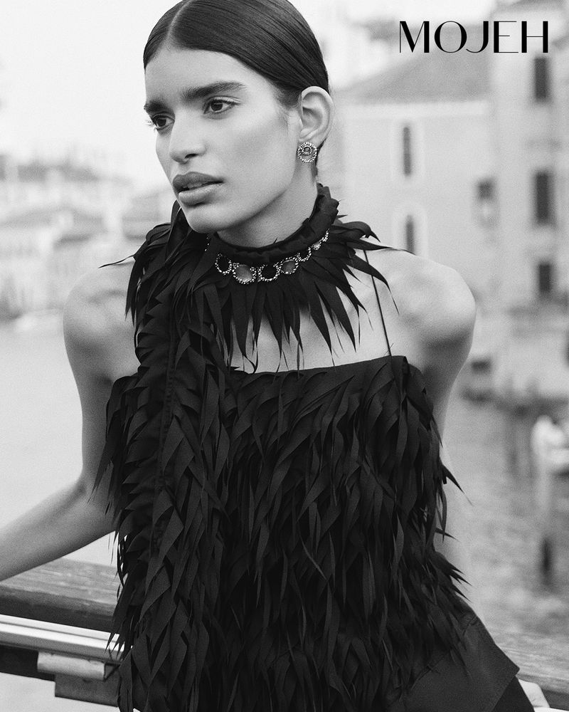 Portrait of an adult woman wearing a black feather-textured top and necklace, buildings in the background, monotone photography.