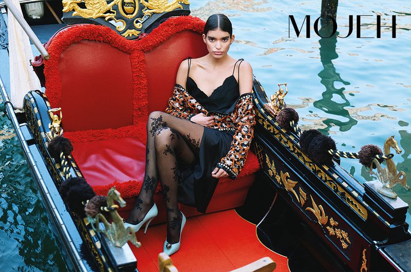 Fashionable adult woman poses in a gondola on a canal in Venice, Italy wearing patterned blouse, black dress, and lace patterned tights.