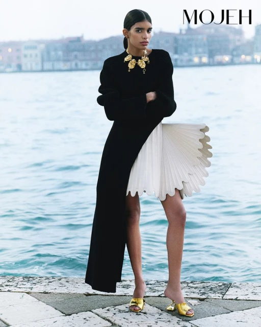 Fashion editorial image of a female model in a black dress and statement necklace, posing outdoors near a body of water.
