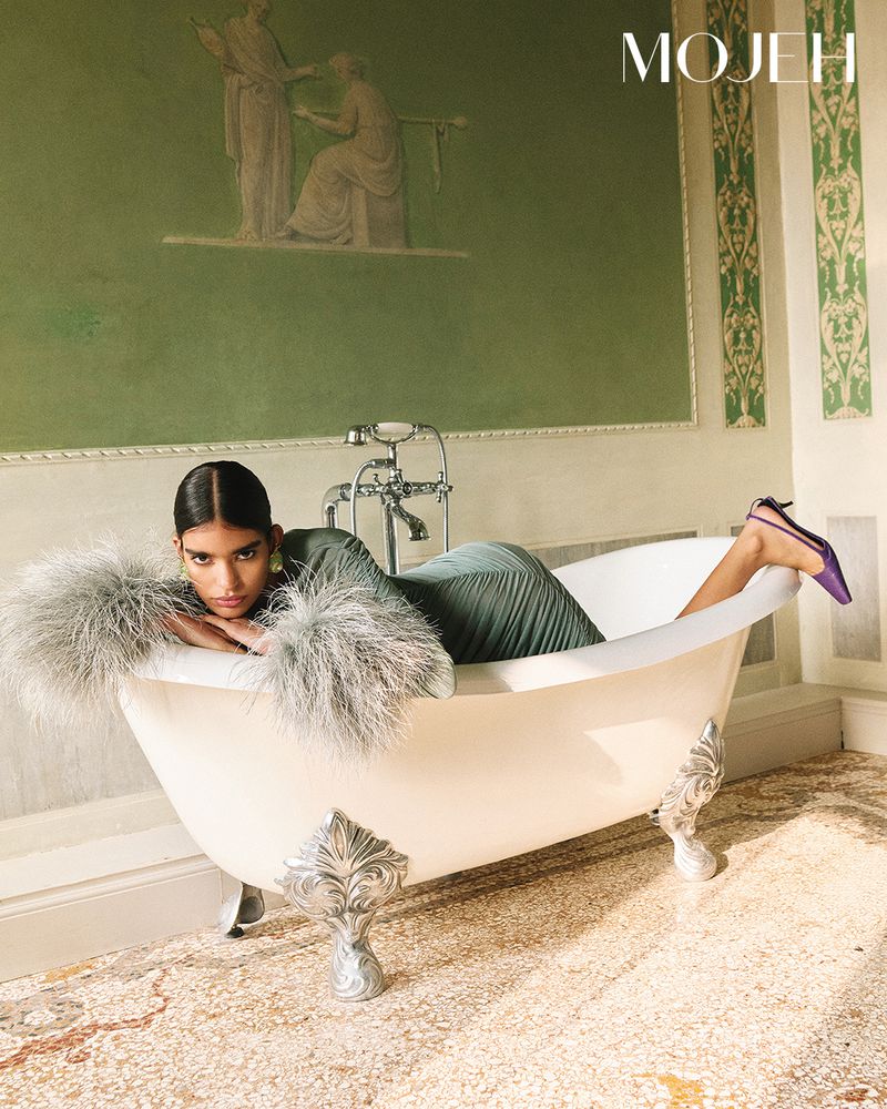 Fashion shot of a woman reclining in a clawfoot bathtub, wearing a dress and fur stole, with a mural in the background.