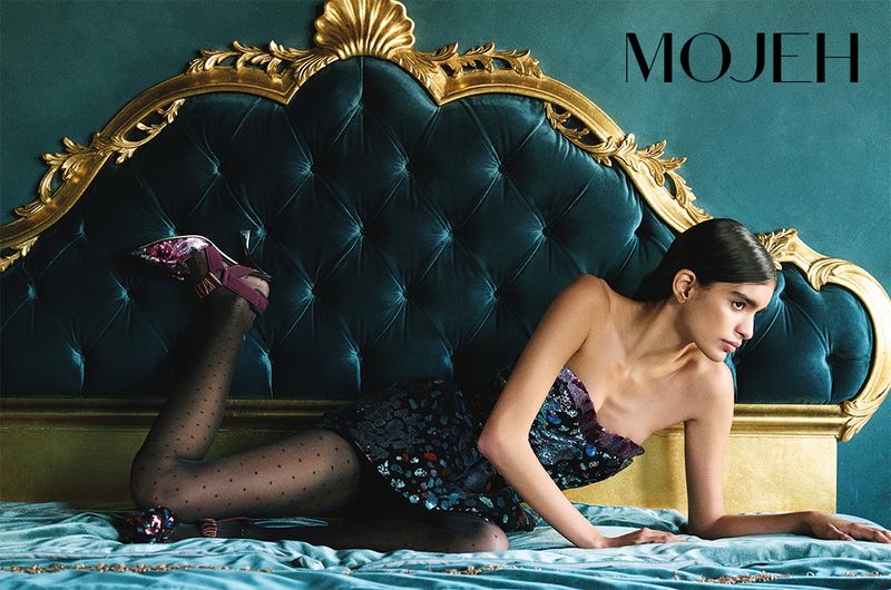 A woman in a dress and heels is lying on a tufted headboard and light blue bedding in an indoor setting with the brand MOJEH in the upper right.